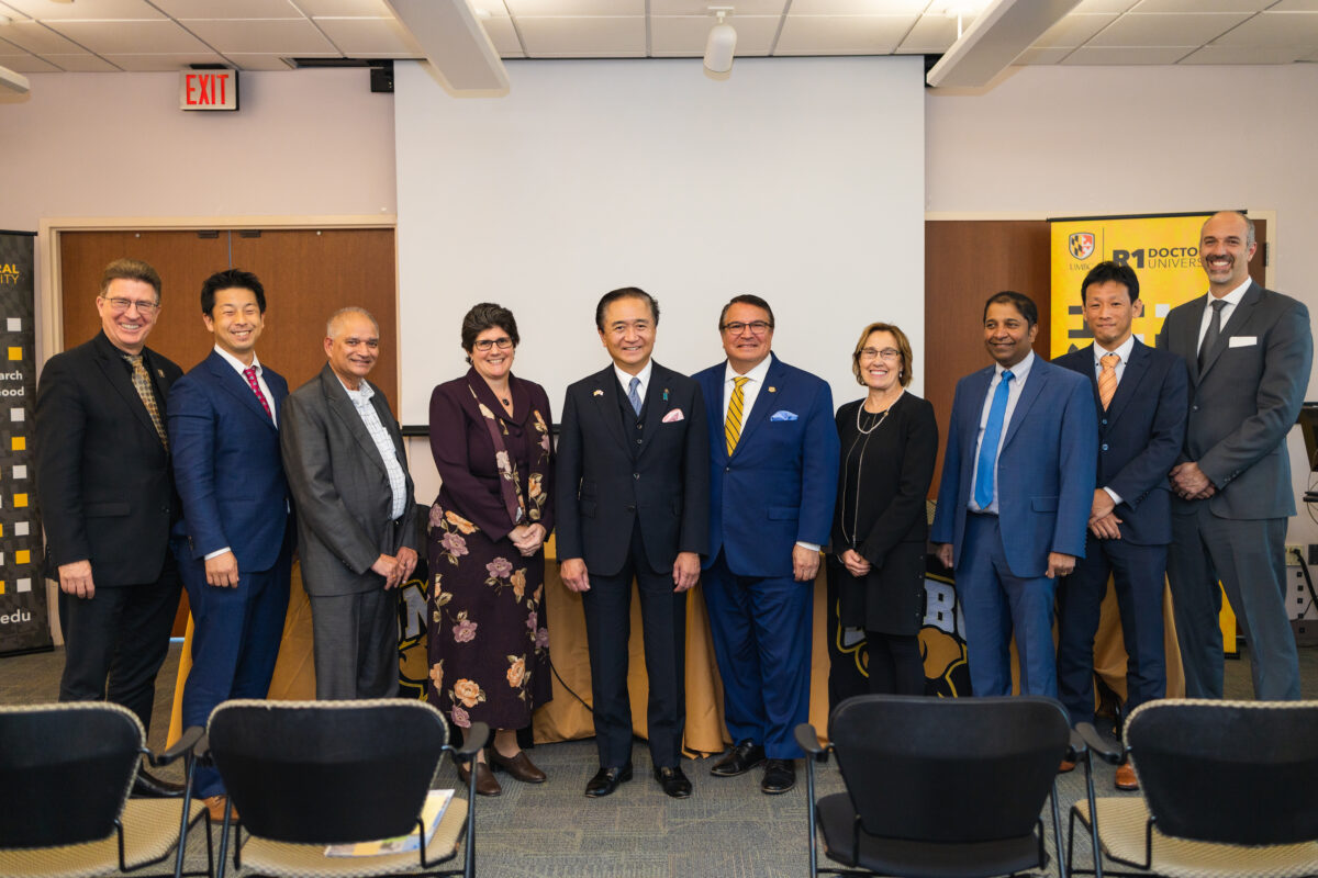 A group of ten people wearing business clothing stand close together smiling at the camera with a blank white projection screen and sign reading, "UMBC R1 Doctoral University" behind them.