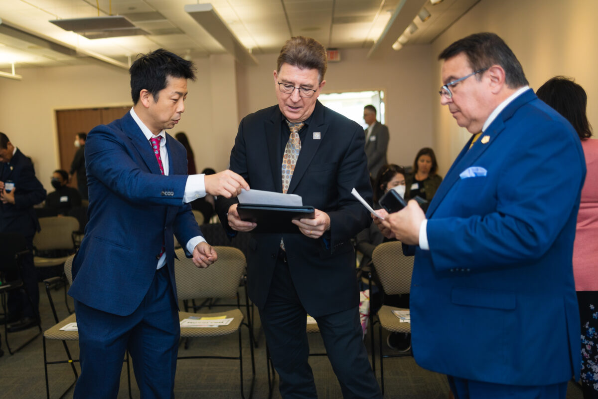 Three people wearing business suits stand next to each other in a room full of people reviewing papers for an event on health aging research and policy.
