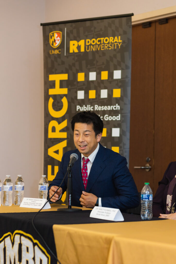 An adult wearing a navy business suit with a red tie sits at a table speaking into a microphone in the background is a black, gold, and white banner with the words UMBC R1 Doctoral University.