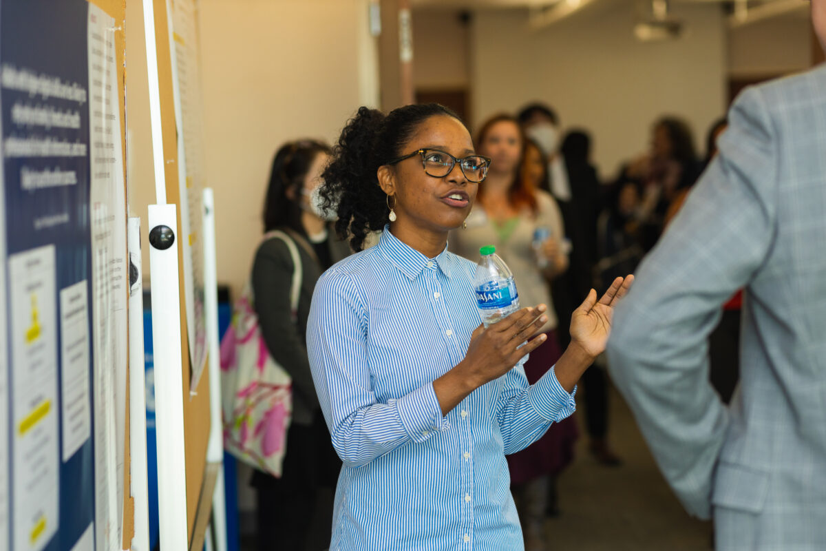 A person with dark black hair pulled back into a ponytail, wearing dark-rimmed glasses, white tear-drop earrings, and a blue and white pin-stripped dress shirt speaks in front of an academic research poster while holding a plastic Dasani water bottle.