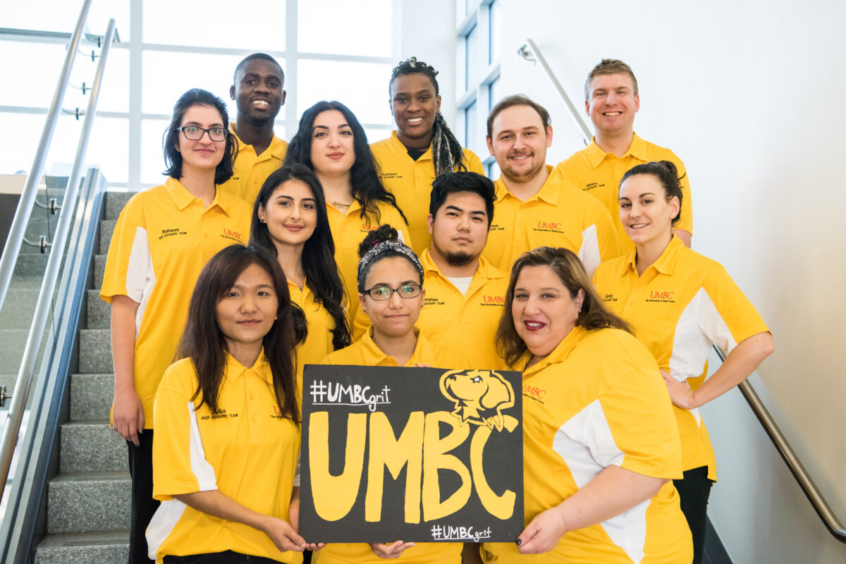 A group of students in gold hold a sign that says @umbcgrit