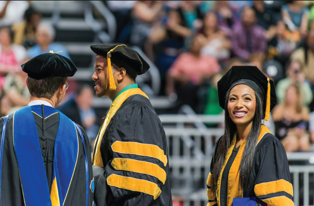 Two black doctoral students smiling on stage during graduation ceremony with UMBC faculty.