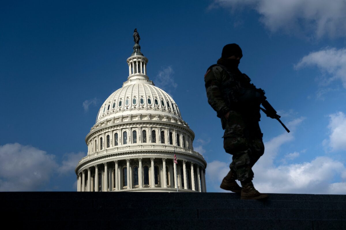 A soldier with a machine gun stands in the shadow with the U.S. Capitol building in the background.