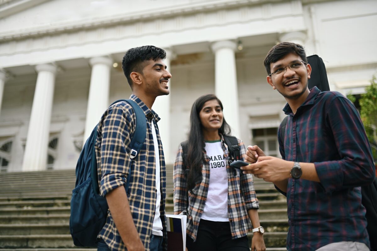 A group of three students wearing backpacks stand close together smiling in front of a building. College enrollment.