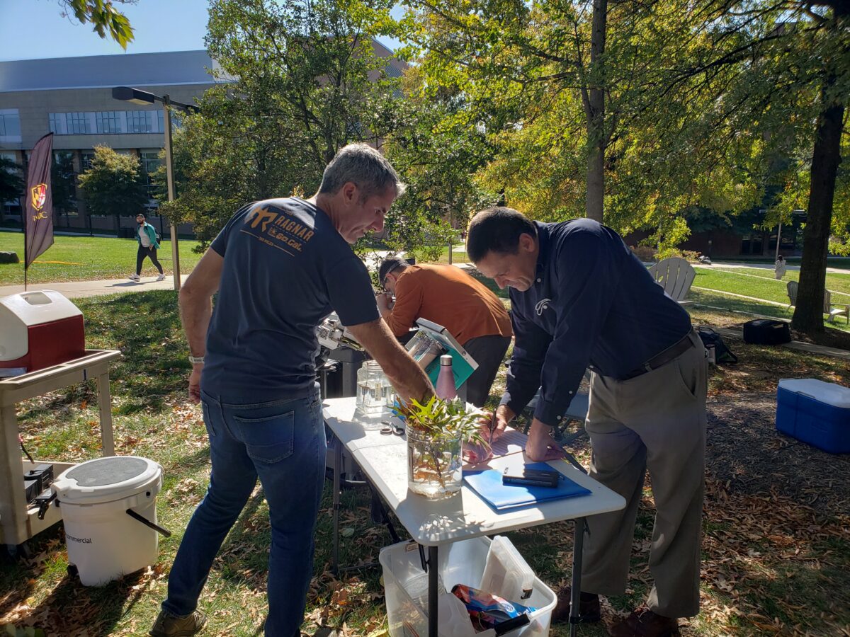 Two researchers stand outdoors over a folding table. One is taking notes, a second pointing to the first's notepad. A tree clipping sits in a glass jar on the table. In the background, another researcher peers into an instrument. The sun is shining.