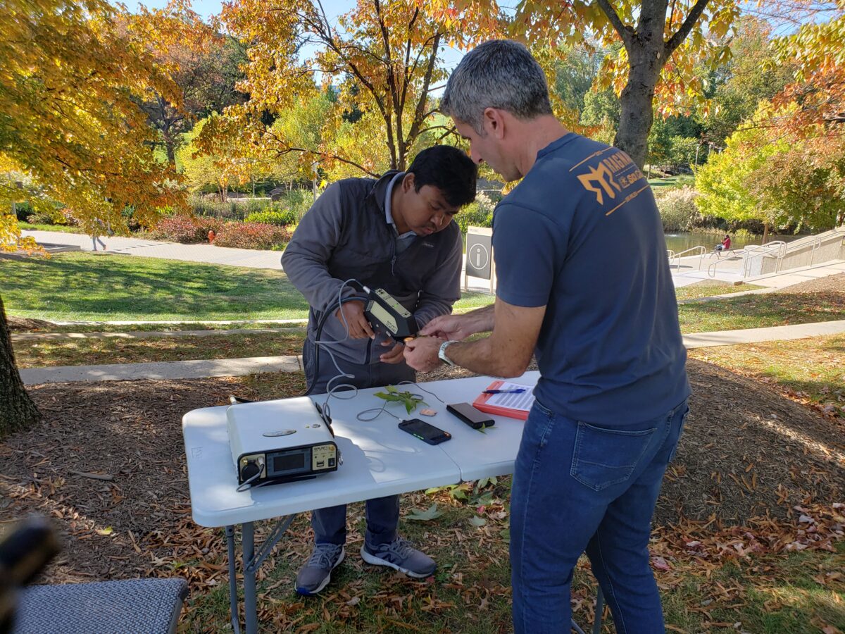 Two researchers stand on opposite sides of a folding table. One holds a leaf, another points an instrument at it to take a measurement. Other equipment lies on the table. Fall foliage in the background.
