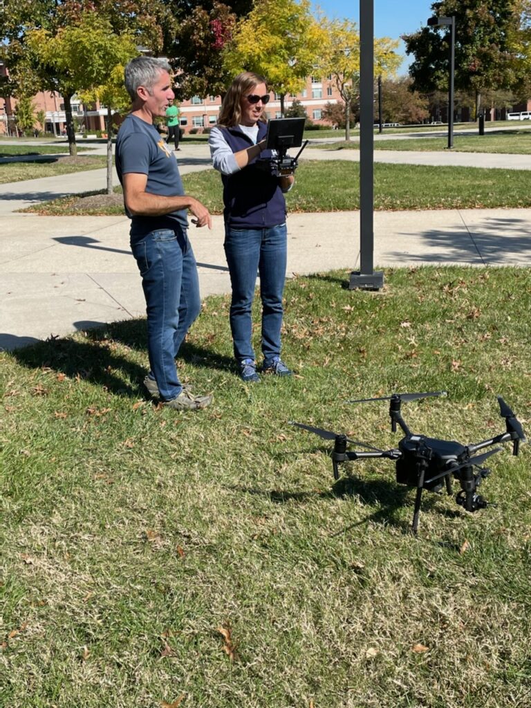 Two people stand outdoors, one holding a tablet-like controller. A quadcopter drone sits on the grass a few feet away.