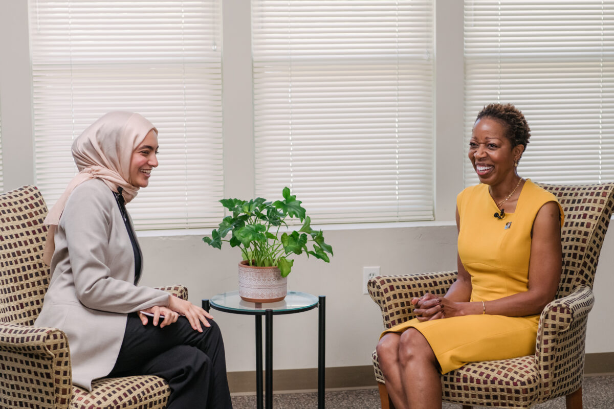 In an office hour set up, two people sit facing each other, engaged in conversation. The student wears a peach headscarf and gray blazer and President Sheares Ashby wears a bright gold dress.