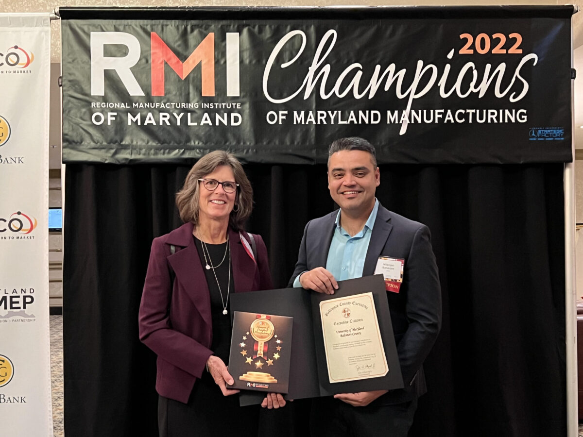 Two smiling people stand, holding awards, in front of a sign that reads, "RMI Regional Manufacturing Institute of Maryland Champions of Maryland Manufacturing 2022"