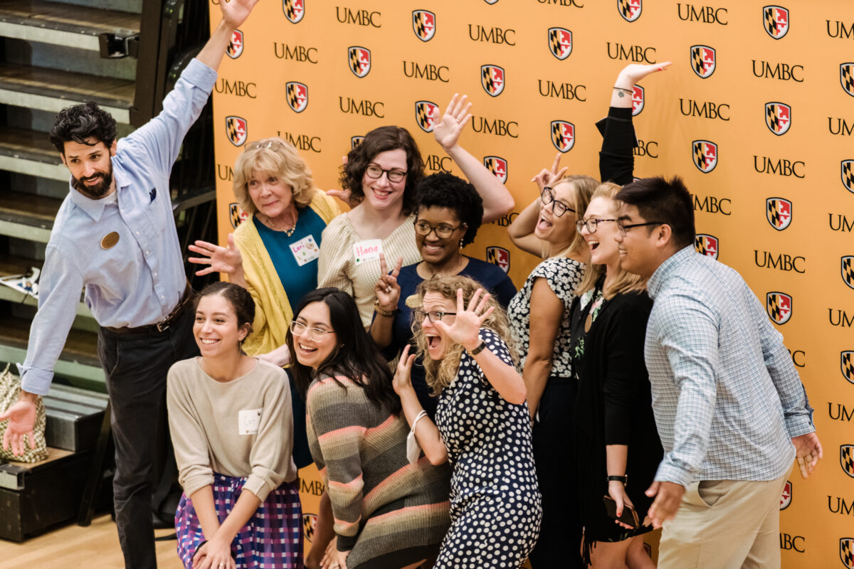 10 adults pose for a group photo, smiling and waving their hands excitedly, in front of a background reading "UMBC"