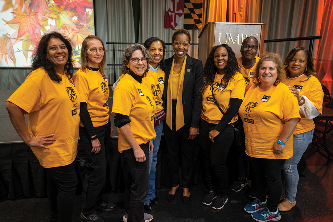 8 women in matching gold t-shirts that read UMBC Family stand in a cluster with the UMBC President in the center.
