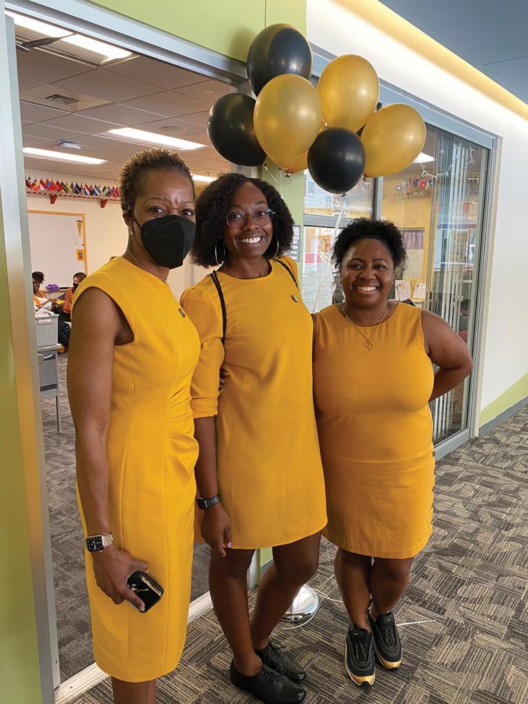 Three women in the same color gold dress stand below an array of black and gold balloons.