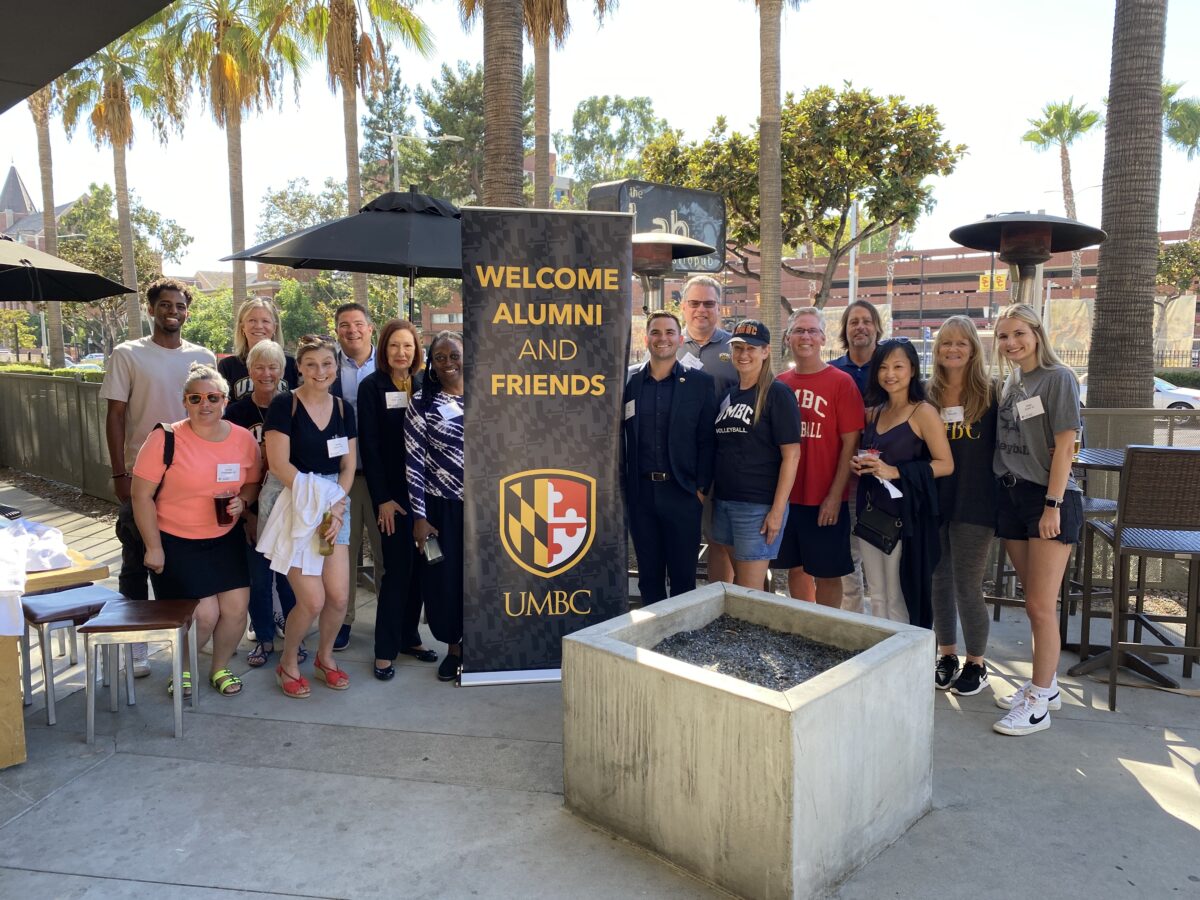 with palm trees in the background, a group of folks stand around a sign that says Welcome UMBC Alumni and Friends