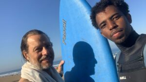 two men stand next to a surf board at the beach
