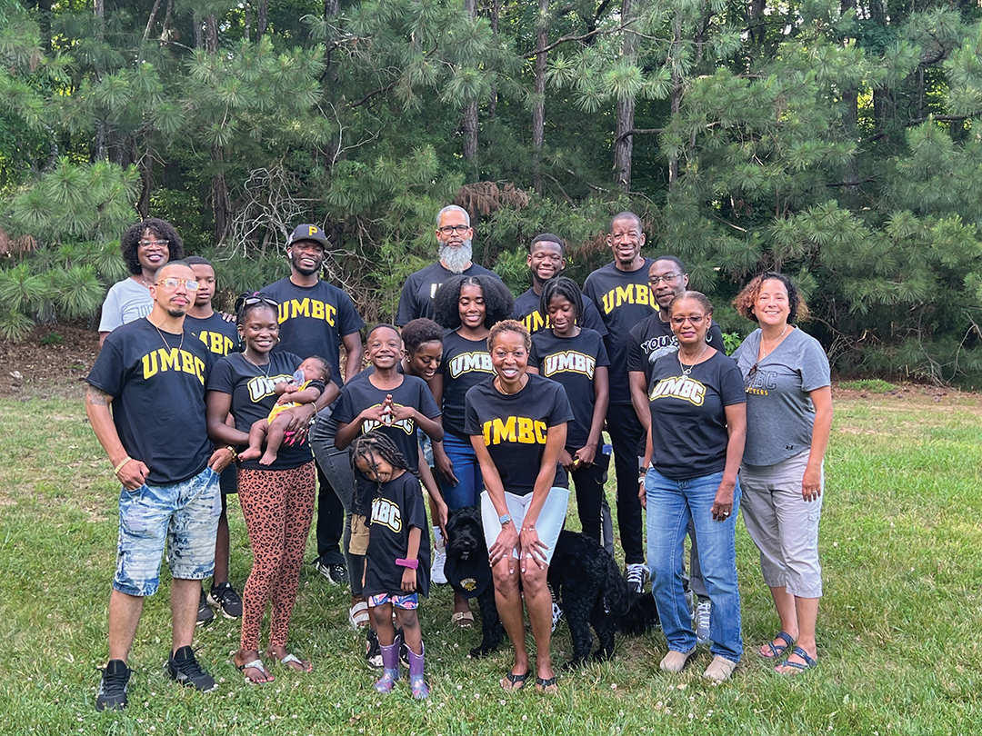 A family of 18 all wear UMBC branded clothing, standing in front of a treeline.