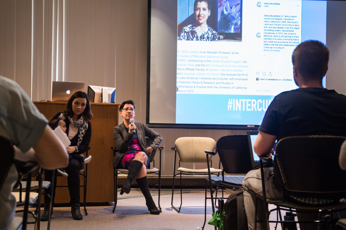 A presenter speaks in front of a projected screen to a small crowd. The talk is titled Intercultural Tales: Learning with Maryland’s Immigrant Communities"