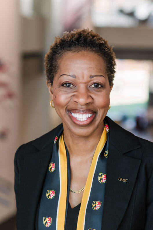 Headshot of President Sheares Ashby. She wears a black jacket with a UMBC pin and a black and gold scarf with the UMBC shield printed on it