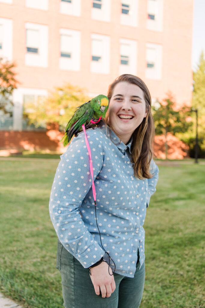 Elle Kreiner with Chicken the parrot (mostly green bird with a yellow crown on his head) stand in front of the public policy building