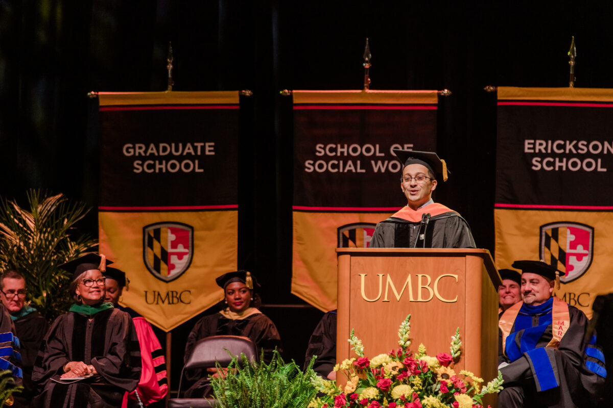 Man in glasses speaking at UMBC podium in graduation regalia