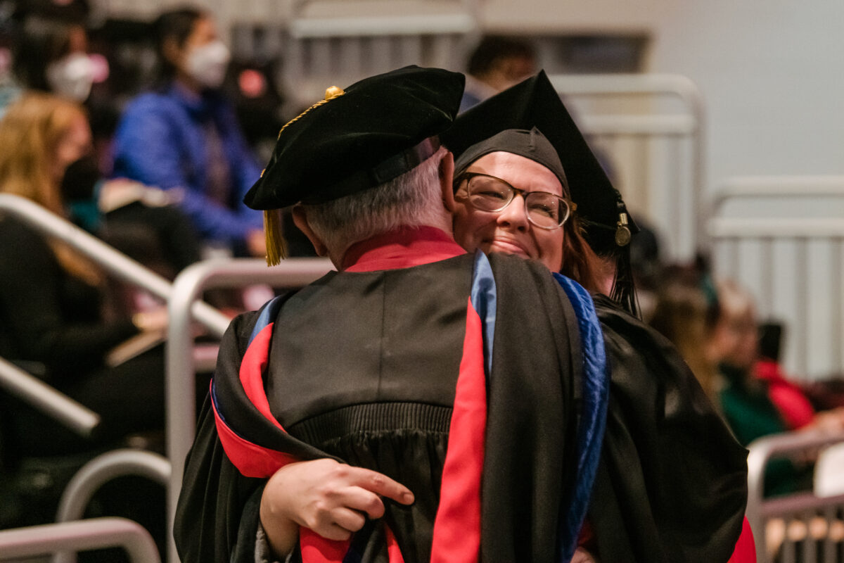 A graduate with glasses hugs a professor in regalia smiling