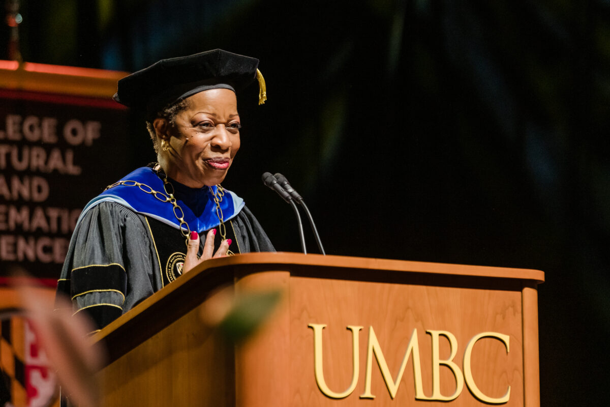 UMBC president stands at UMBC podium in graduation regalia with her hand over her heart.