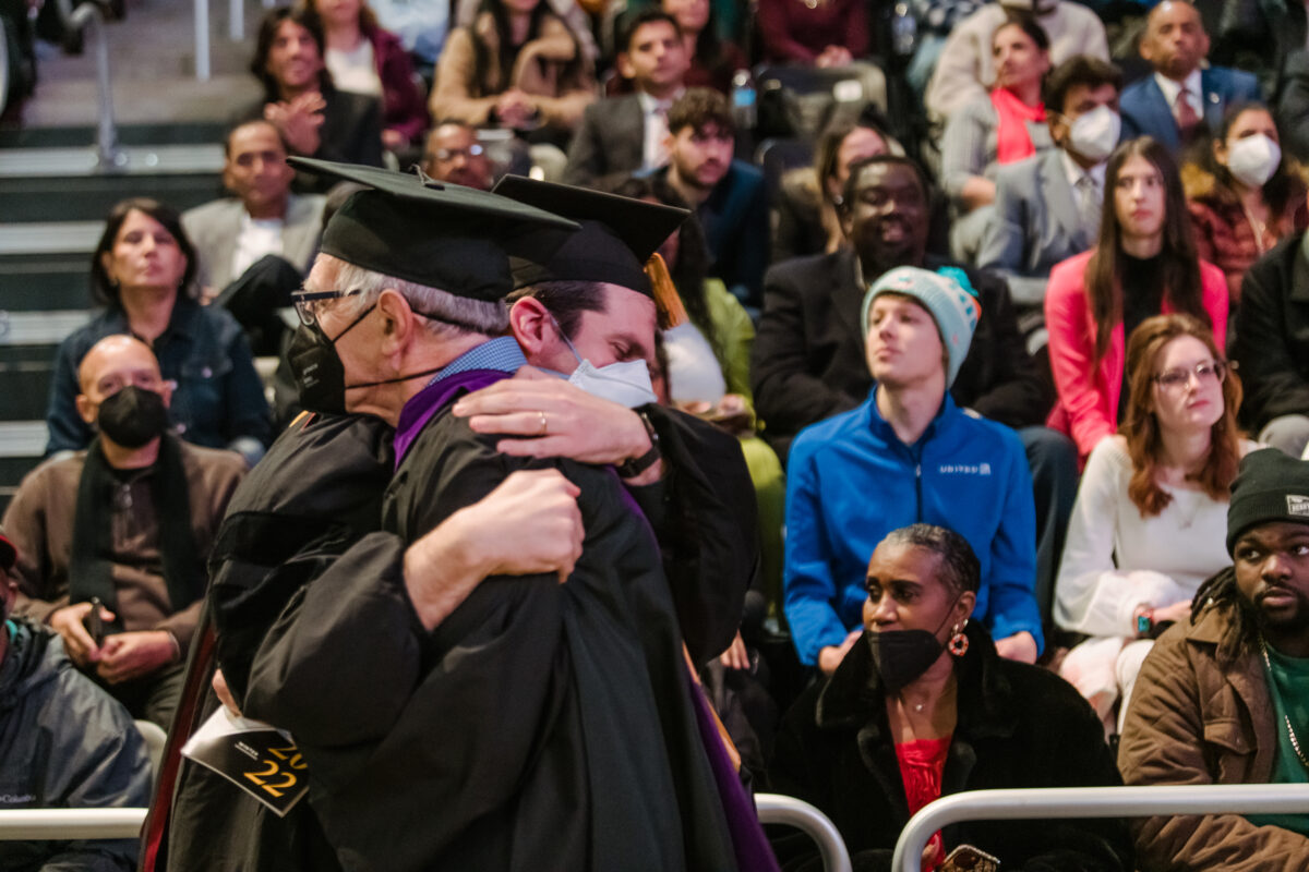 A white-haired gentleman hugs his son in the audience, both wearing graduation regalia.