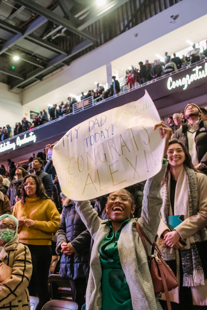 Person smiling widely holding a sign in the audience that says "My dad graduates today! Congrats, Alex!"