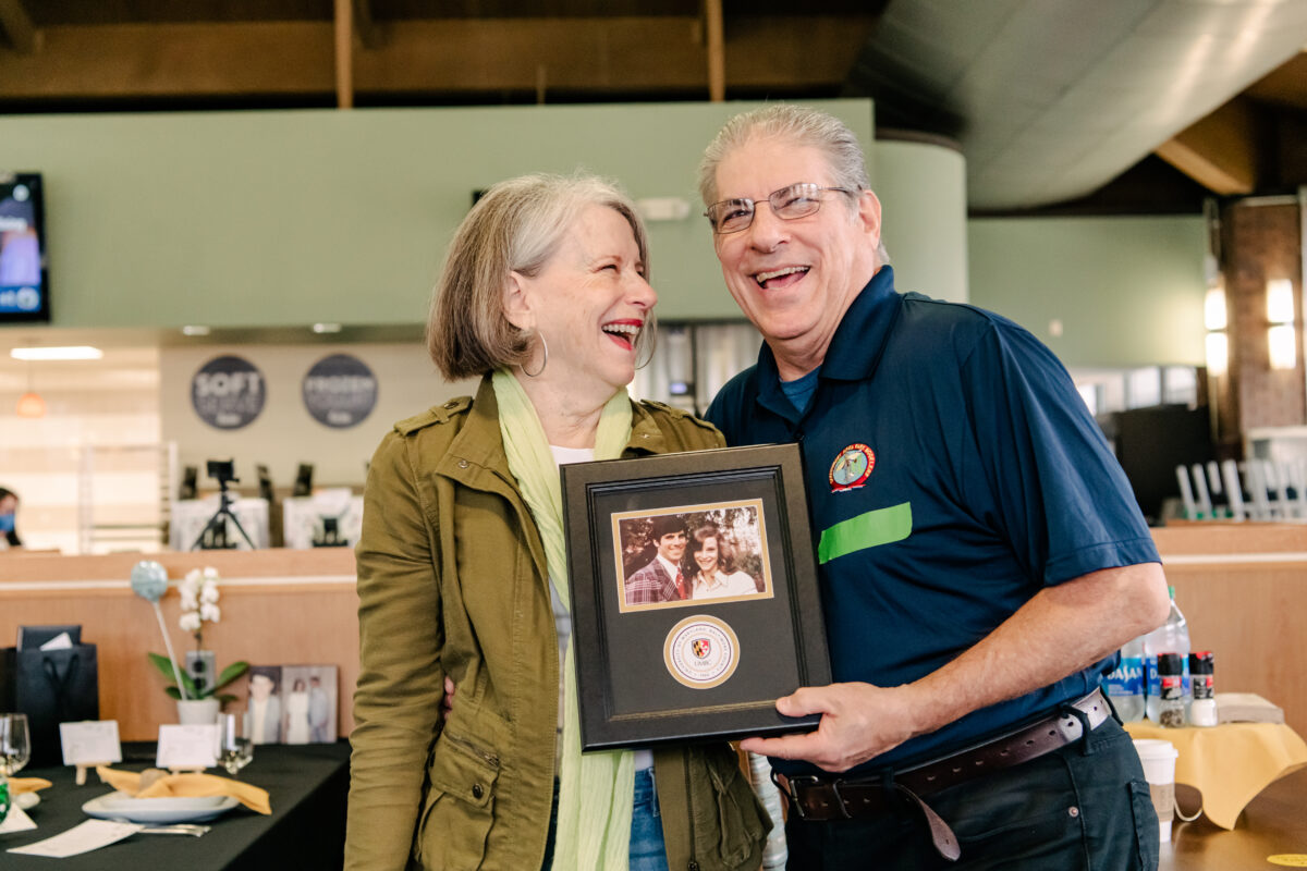 A man and a woman smile and laugh at one another while holding an aged photograph of themselves.