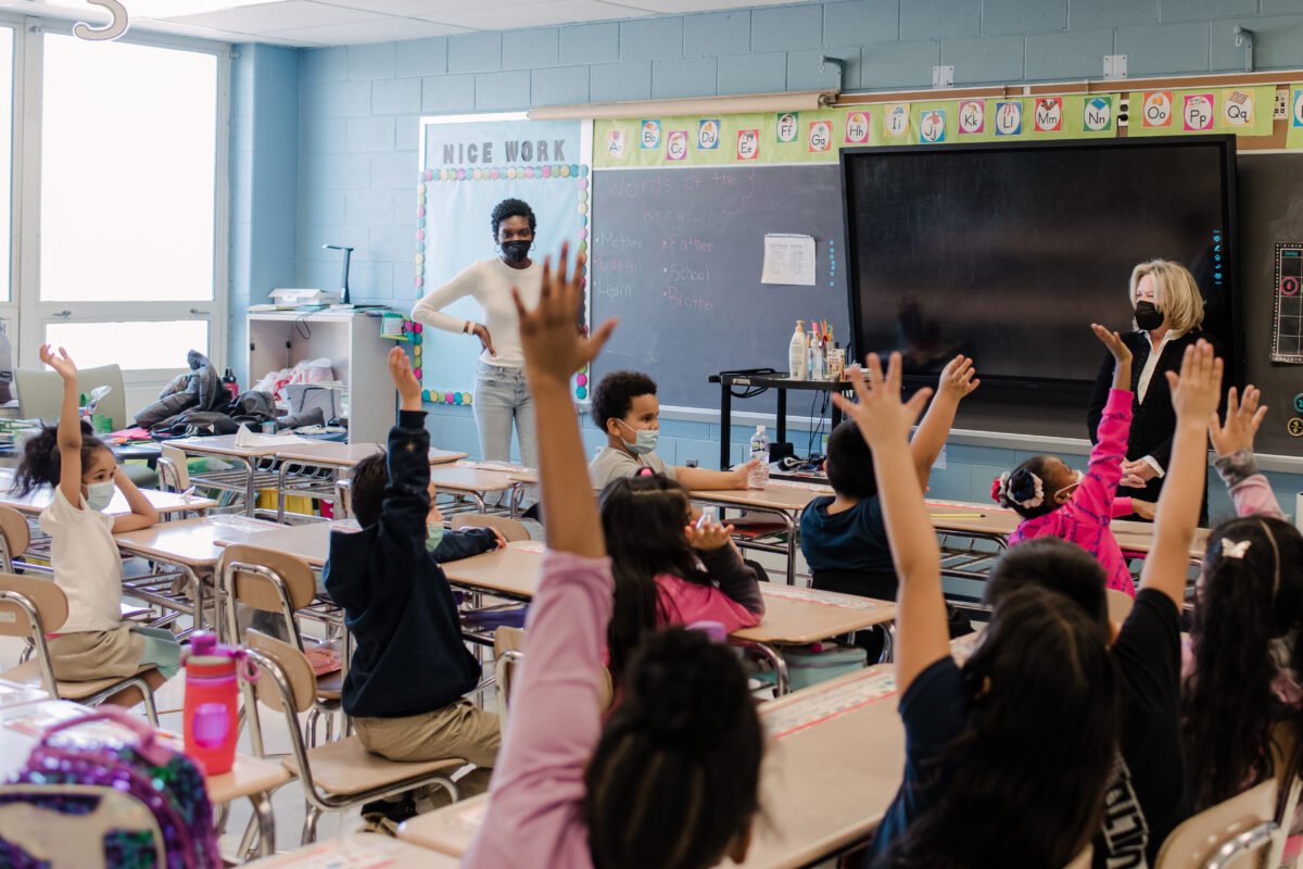 A group of young students raise their hands energetically in a classroom while two women look on.