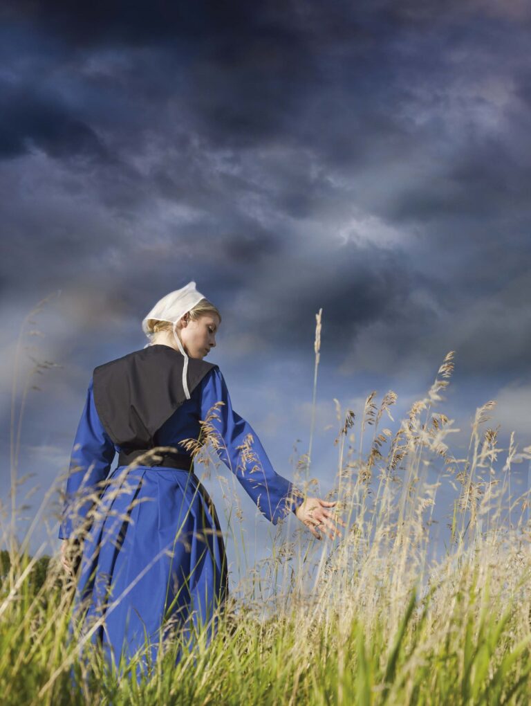 An Amish woman in a blue and black dress and white hat stands in a field against a dark sky