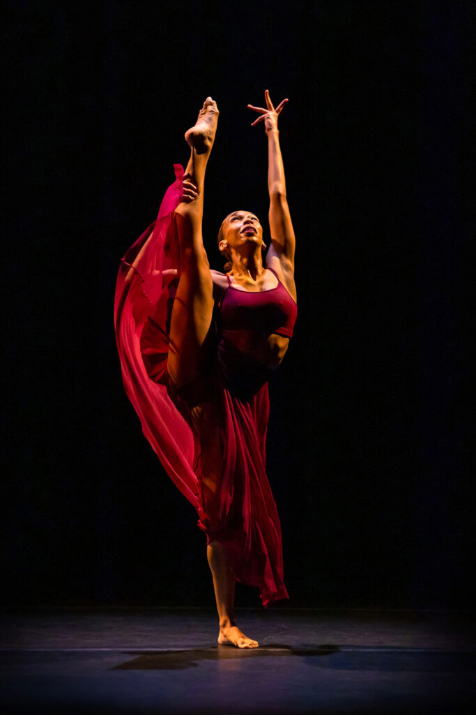A Black woman in a red costume dances on stage, with one leg lifted high over her head