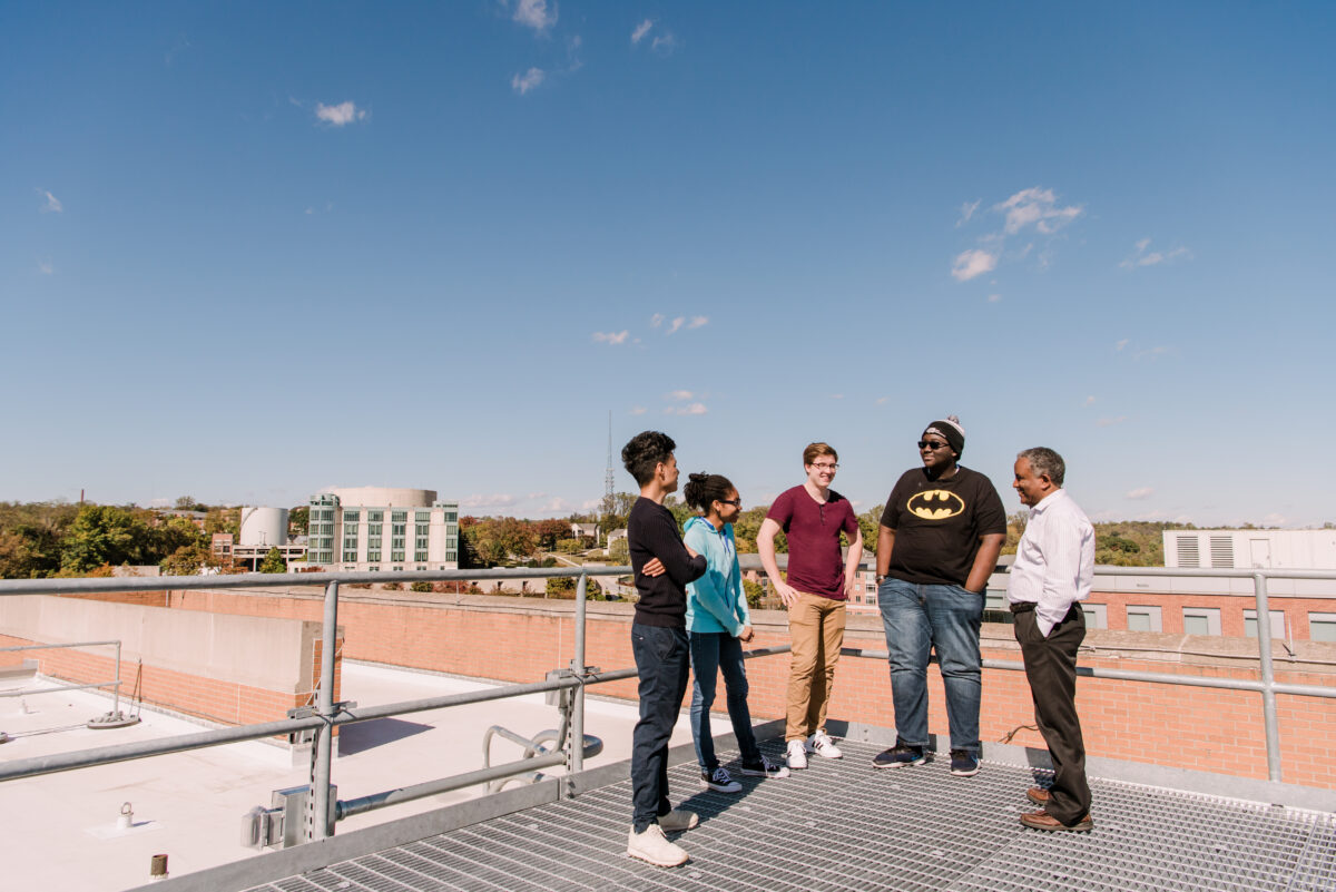 five people stand on a roof, backed by wide blue sky and a distant view of the UMBC Library.