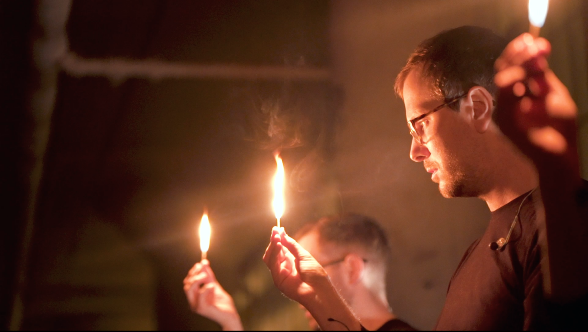 A man with glasses looks down at the flame from a candle, and other people near him seem to do the same, although they are out of focus