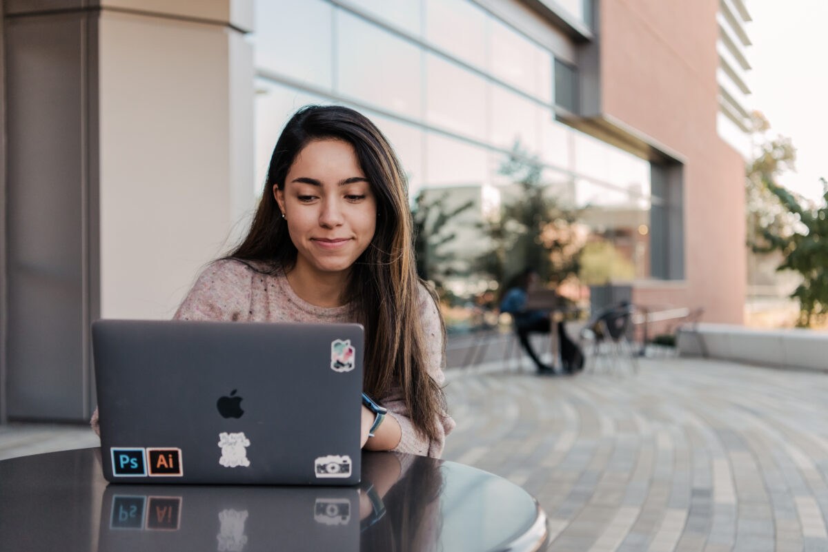 A female student with long brown hair works outside at a table on a laptop.