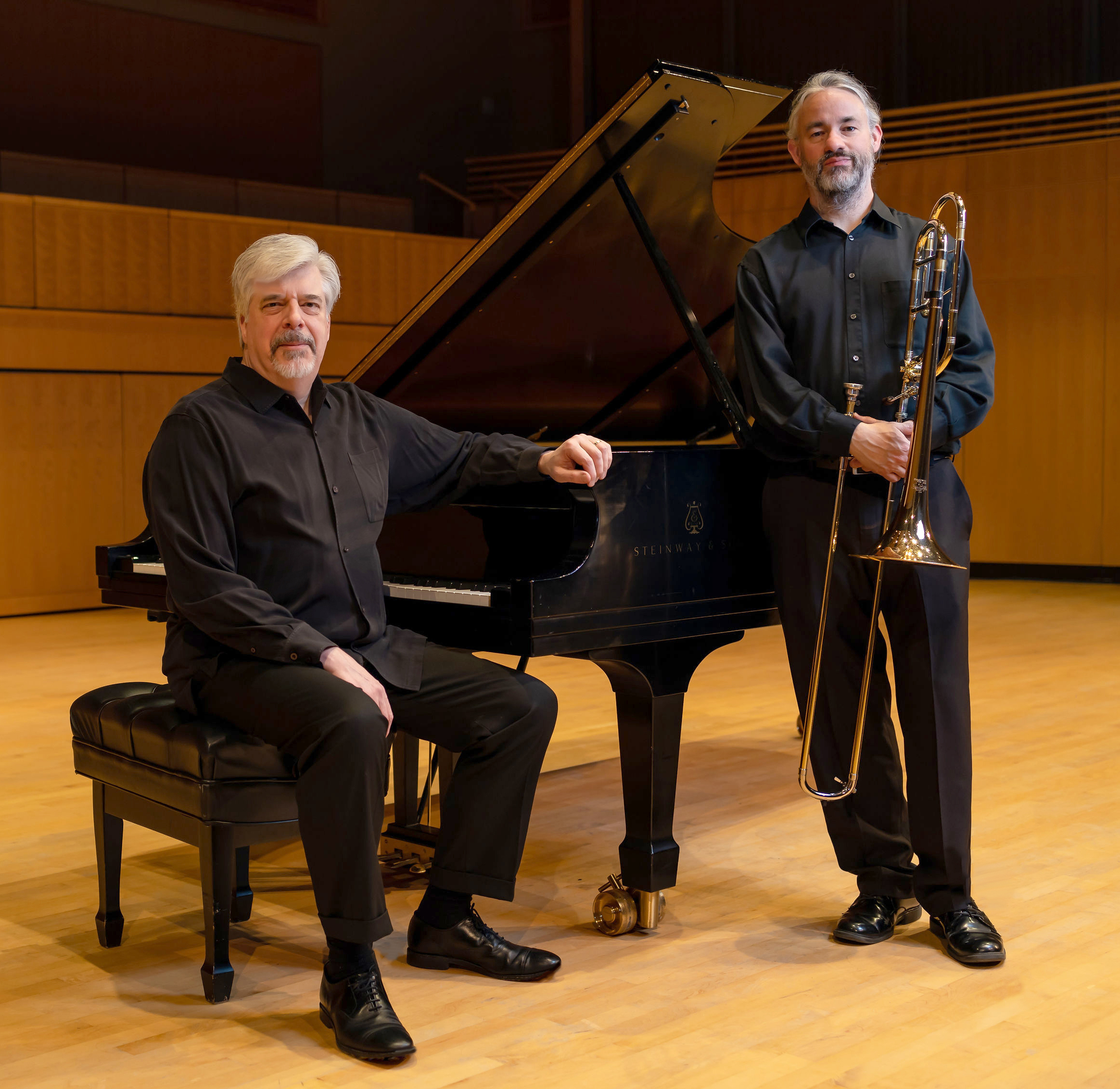 On a concert stage, two white men with gray hair and beards, the left one sitting at a piano the second standing and holding a trombone