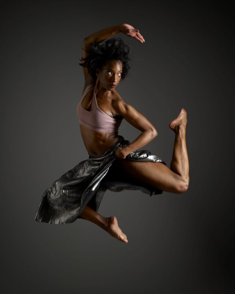 A female Black dancer leaps into the air in front of a dark background, wearing a pink top and black skirt
