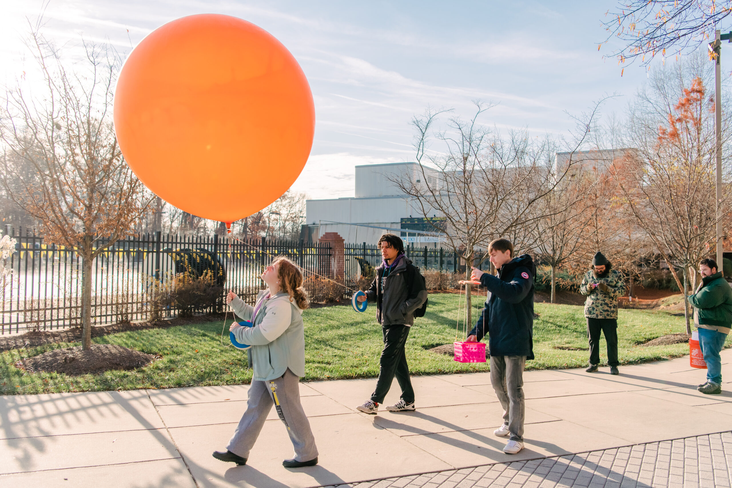 Three student walk down a sidewalk on campus holding an orange balloon about five feet in diameter tethered by ropes a few feet above their heads.