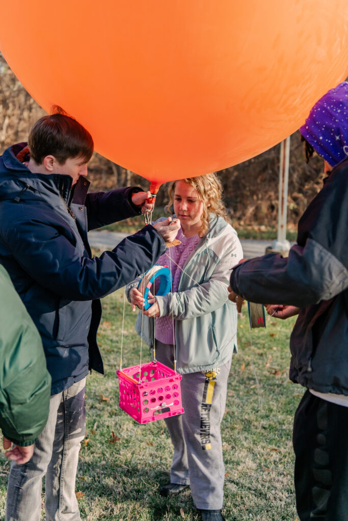 Three students stand directly underneath a large orange balloon; a pink crate dangles from the bottom of it, tethered by white strings a few feet long. 