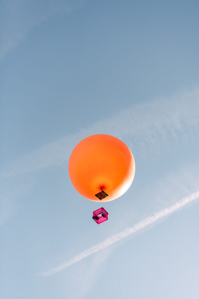 A pink crate dangling from the bottom of a balloon holds a small digital camera horizontal as the balloon flies. 