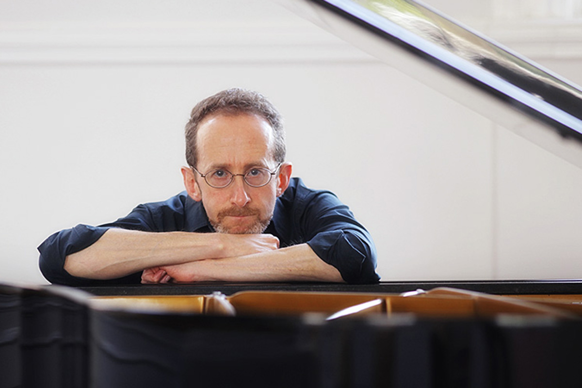 A white man with glasses, brown hair and a goatee rests his head on his arms, which are folded over the top of a piano