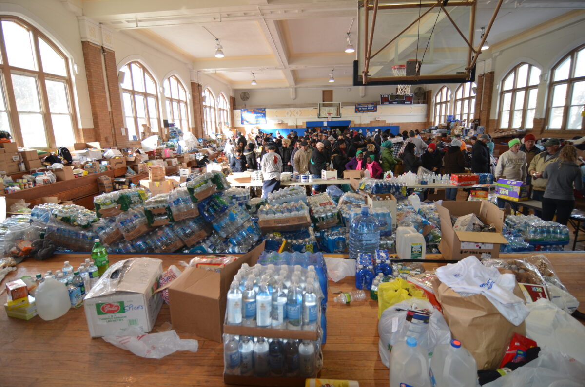 Piles of packaged food, diapers, and bottled water fill a school gymnasium in New York City as a crowd of people wait to receive aid.