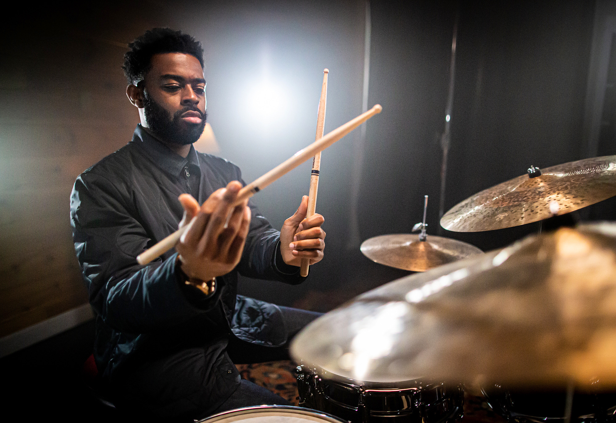 A Black man with a beard, wearing a black top, holds two drumsticks and plays a drum set