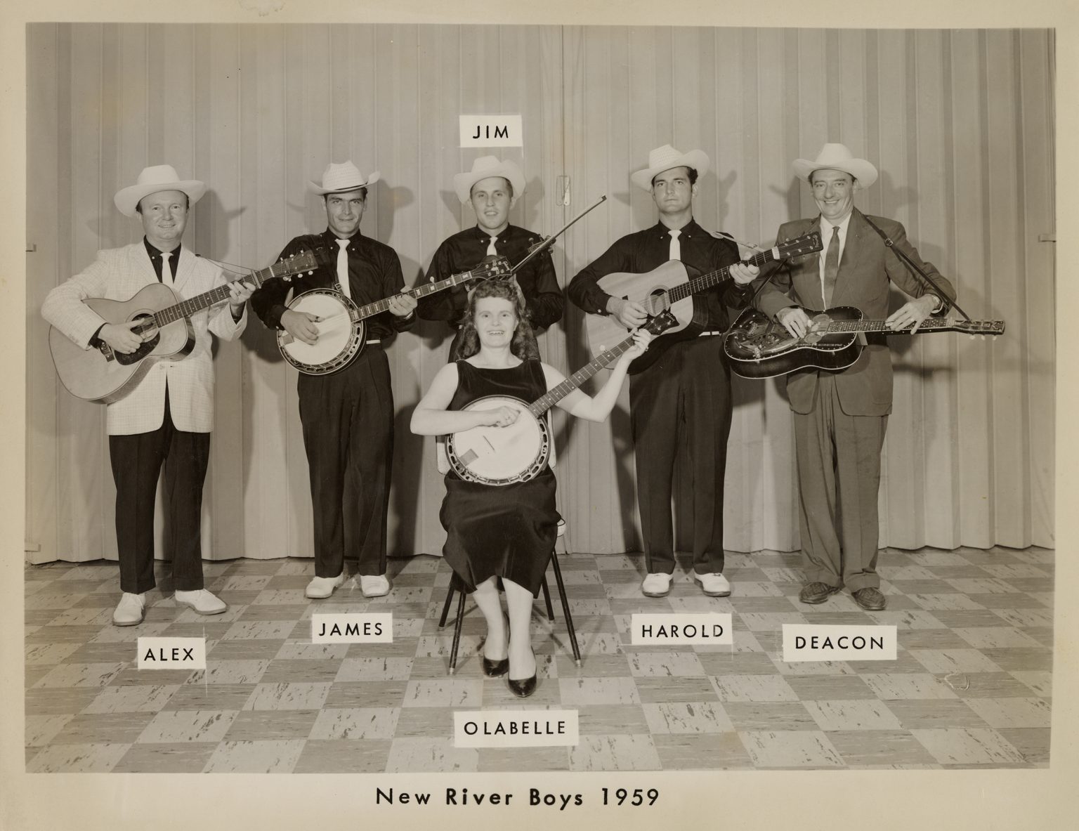 In a sepia toned photograph, five white men with guitars, a banjo and a violin stand behind a seated woman holding a banjo. Each man is identified by their names — Alex, James, Jim, Harold, Deacon — and the woman as Olabelle. A caption at the bottom says New River Boys 1959.