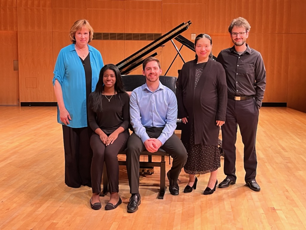 Five people — three women and two men, some standing and some seated — are positioned in front of a grand piano, and they smile at the camera