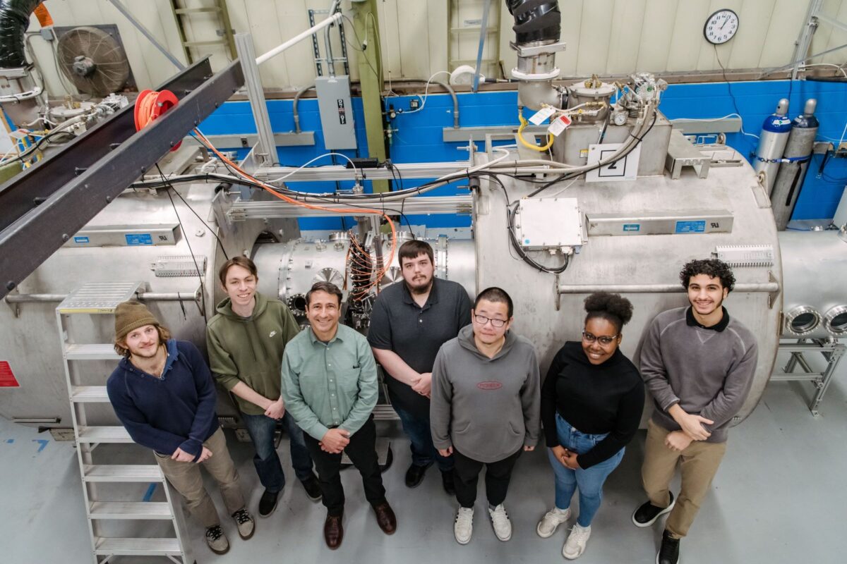 Seven researchers stand in front of a room-sized machine and smile up at the camera.