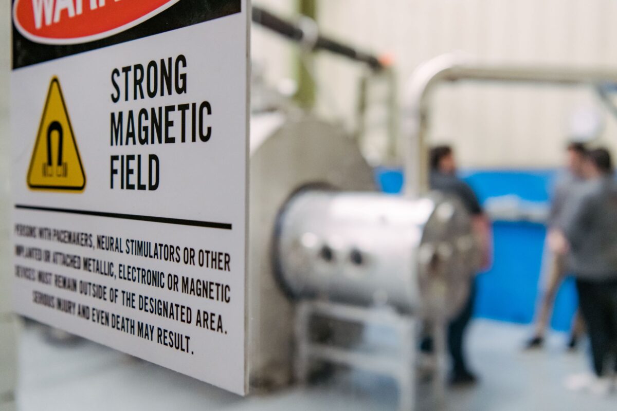 A sign warns in the foreground of a strong magnetic field. Behind the sign is a large machine.