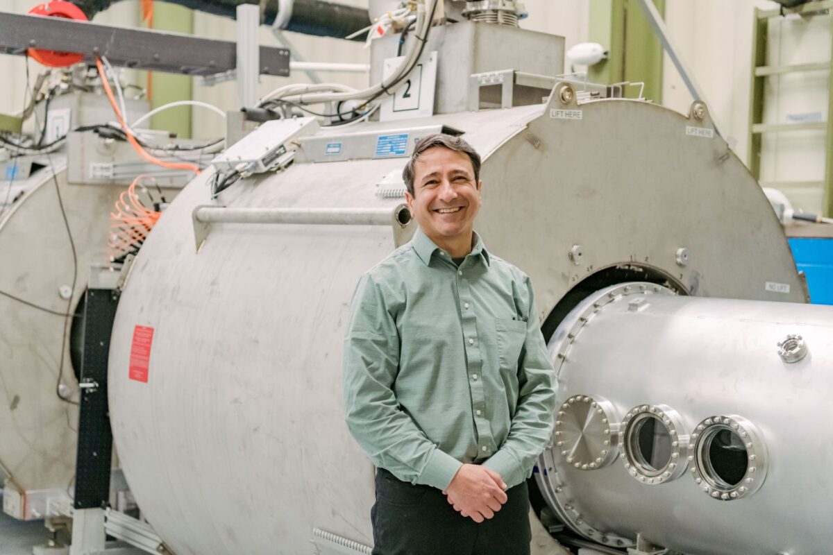 A researcher stands smiling at camera. Large room-sized machines with wires and metal cylinders in background is for testing fusion concepts.