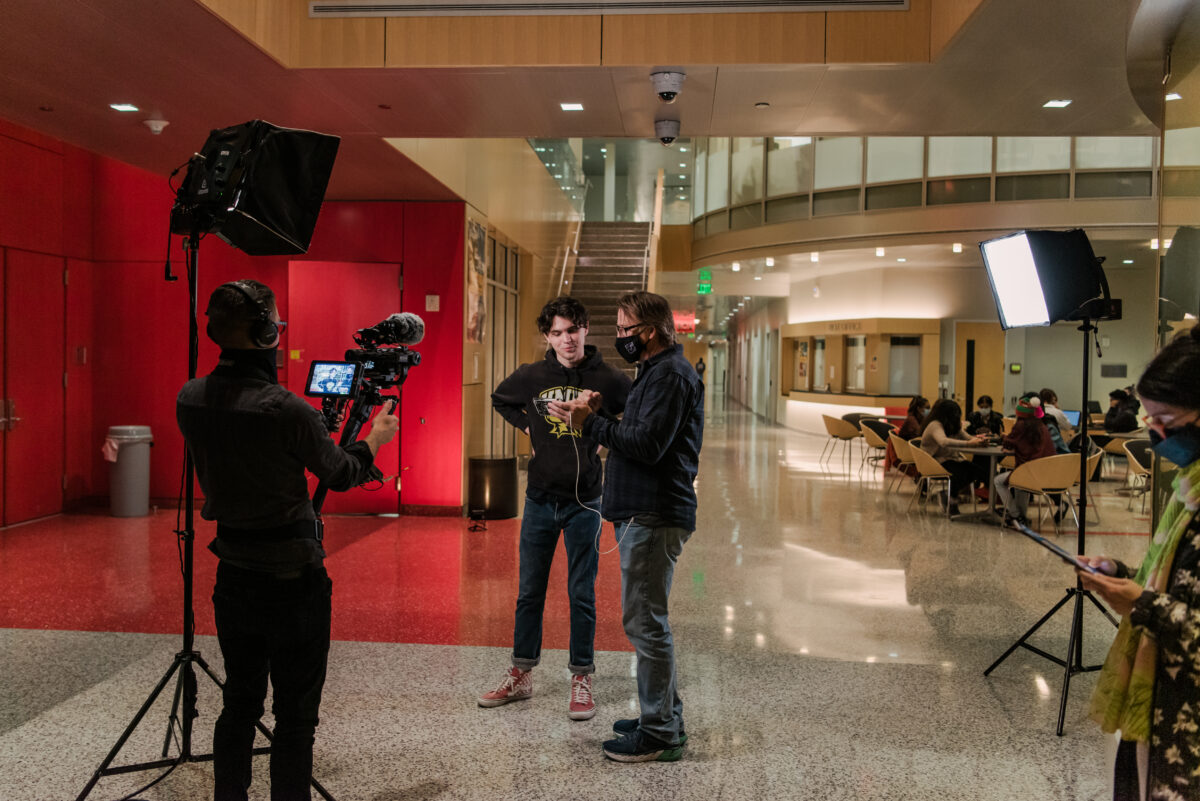 A student stands looking at a phone screen with a man while a camera operator looks on.