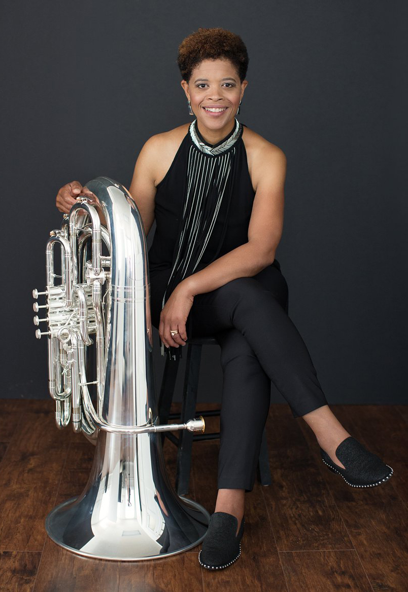 A Black woman, wearing a black top, black pants and black shoes, is seated next to a silver tuba