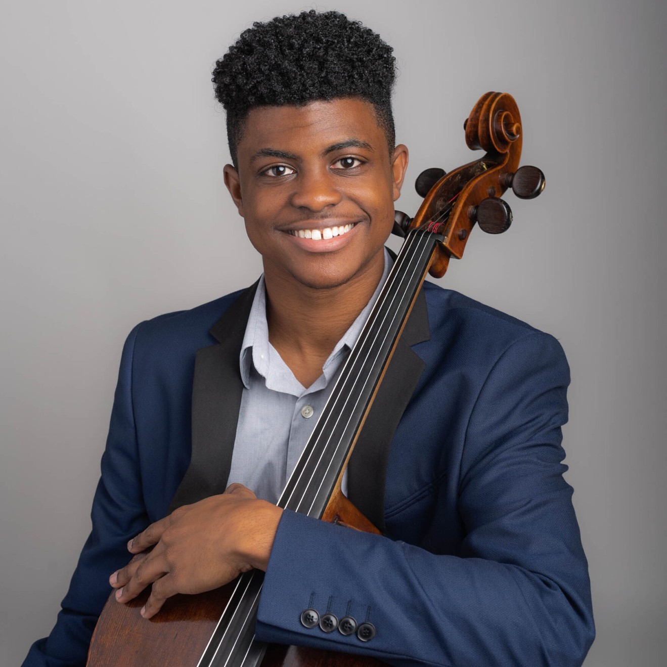 A young Black man dressed in a light blue shirt and dark blue jacket, smiles at the camera while holding a cello